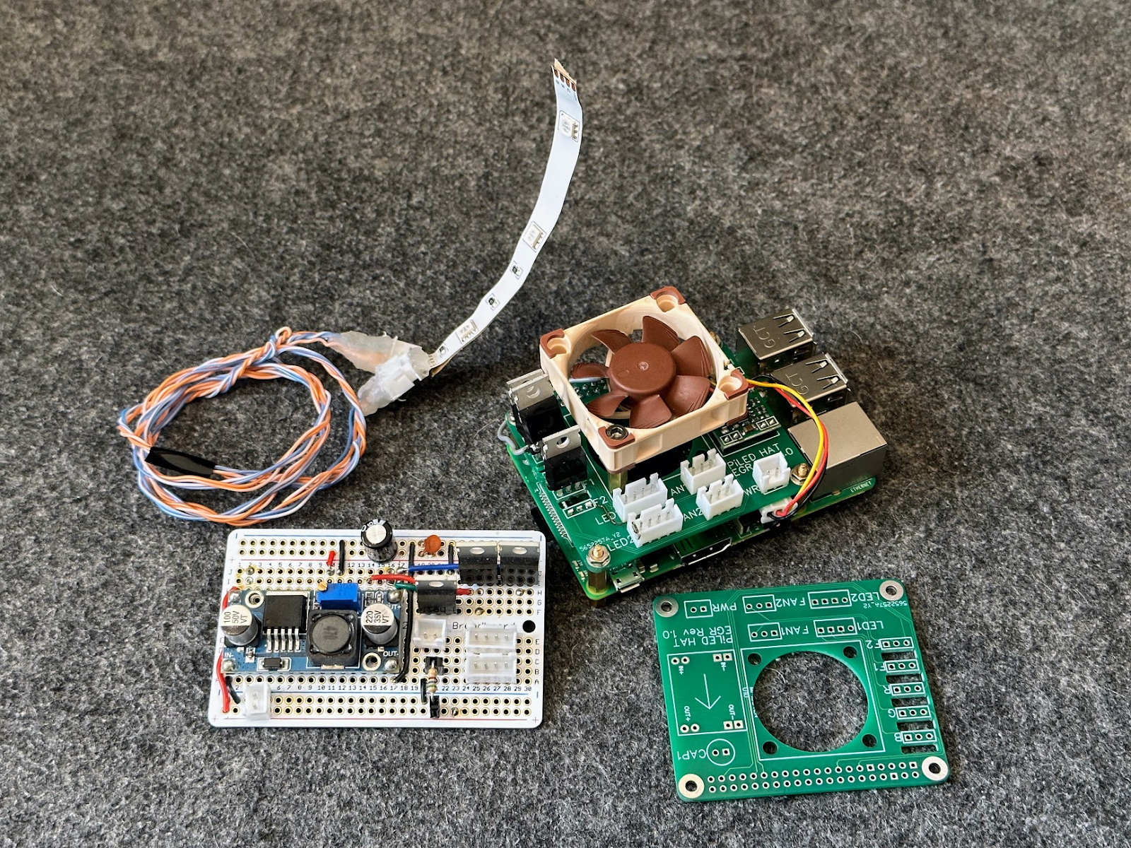 James's photo of the project parts on a grey desk mat: LED strip, twisted orange and blue wires, prototyping board with buck converter, Raspberry Pi with fan, and a bare green PCB.