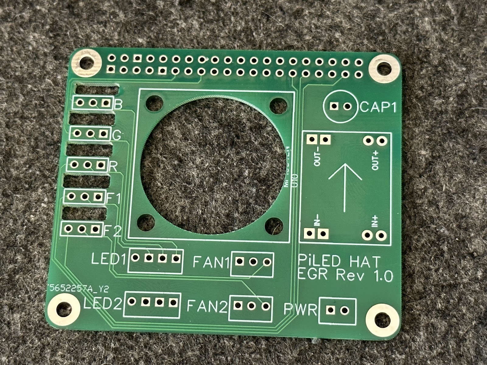 Bare square green PiLED HAT circuit board on grey carpet, with a large central fan opening and white component markings.